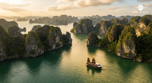 Aerial view of Ha Long Bay limestone karsts emerging from emerald water, golden hour light, traditional junk boat in for