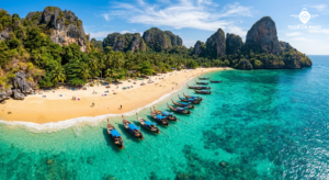 Aerial view of turquoise tropical beach with traditional longtail boats, golden sand, limestone cliffs, bright midday su